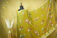 ARN21002 Mustard-green saree features artistic hand-painted floral patterns, a gold zari border, and an intricate traditional elephant-motif pallu.